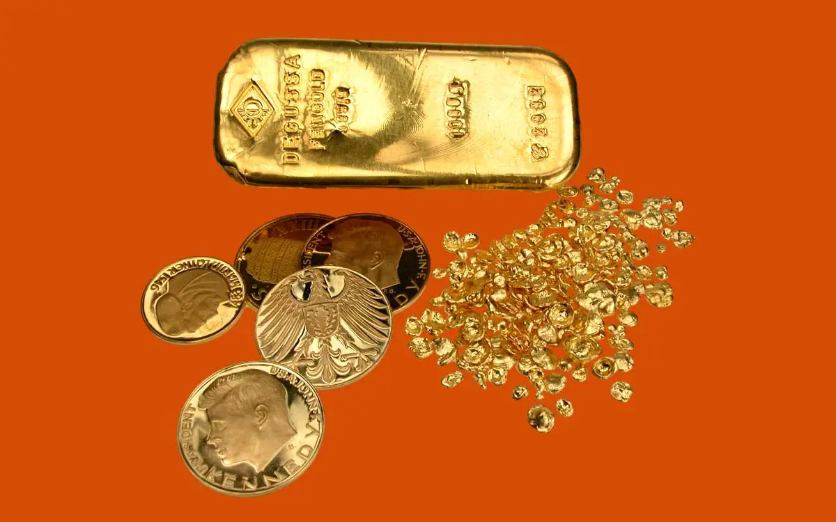 Ein großer Goldbarren von Degussa liegt zusammen mit mehreren Goldmünzen und einem Haufen Gold-Granulat auf einer orangefarbenen Fläche.