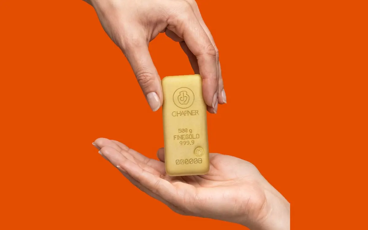 Eine Hand hält einen vertikal ausgerichteten 500g Goldbarren von C. Hafner über eine zweite, geöffnete Handfläche vor einem kräftigen orangefarbenen Hintergrund.