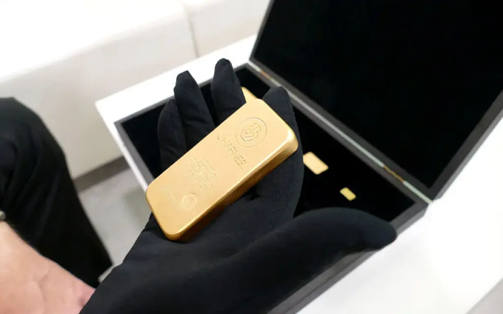 Eine Hand in einem schwarzen Stoffhandschuh präsentiert einen glänzenden Goldbarren von Heimerle + Meule vor einer geöffneten, edlen Schatulle.