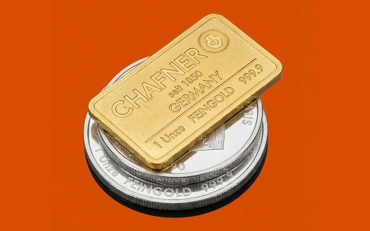 Ein 1-Unzen-Goldbarren von C. Hafner liegt auf zwei gestapelten Silbermünzen vor einem kräftigen orangefarbenen Hintergrund.