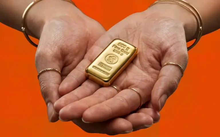 Zwei hohle Handflächen halten einen glänzenden 500g Goldbarren von Heimerle + Meule vor einem kontrastreichen orangefarbenen Hintergrund.
