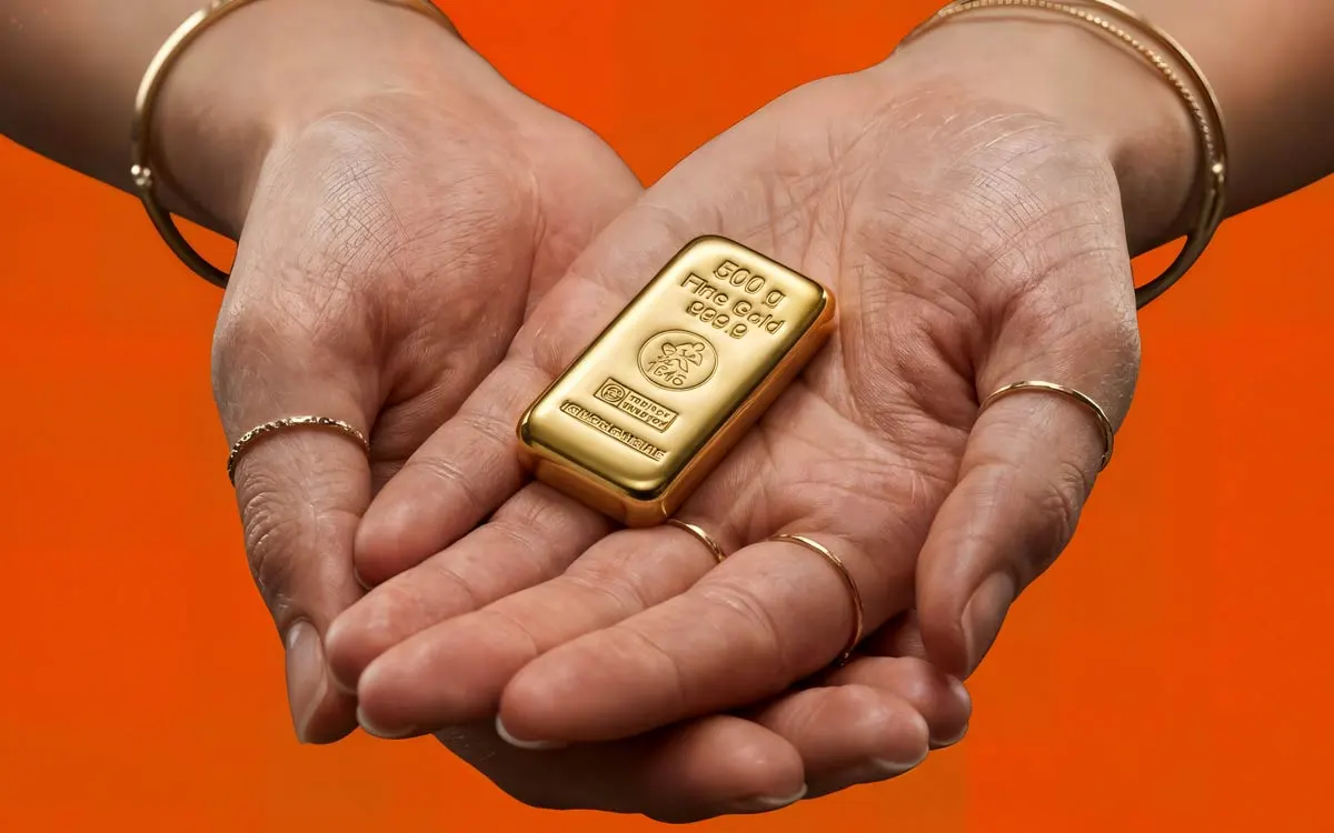 Zwei hohle Handflächen halten einen glänzenden 500g Goldbarren von Heimerle + Meule vor einem kontrastreichen orangefarbenen Hintergrund.