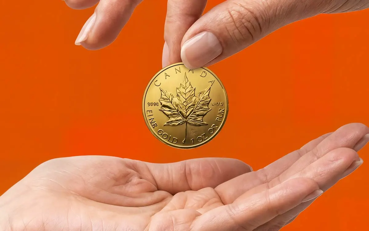 Eine Hand hält eine Maple Leaf Goldmünze über eine geöffnete Handfläche vor einem orangefarbenen Hintergrund.