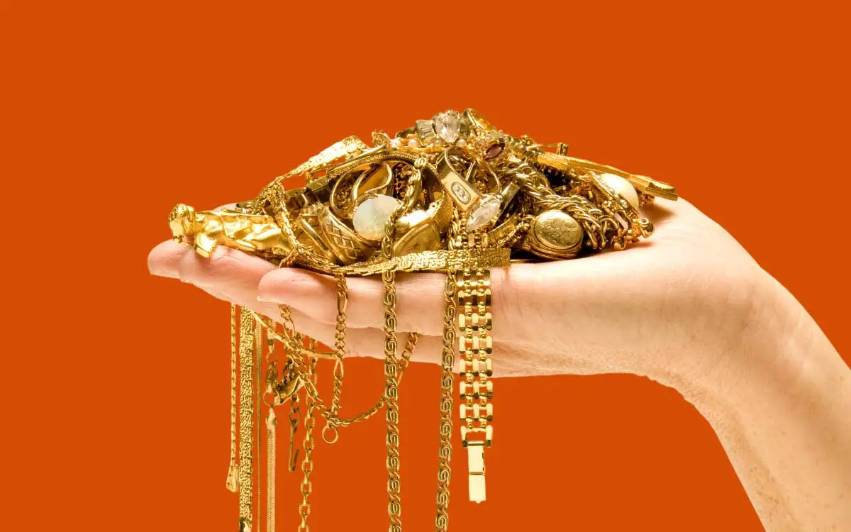 Eine Hand hält eine Vielzahl an Goldschmuck, darunter Ringe, Ketten und Anhänger, die teilweise herabhängen, vor einem kräftigen orangefarbenen Hintergrund.